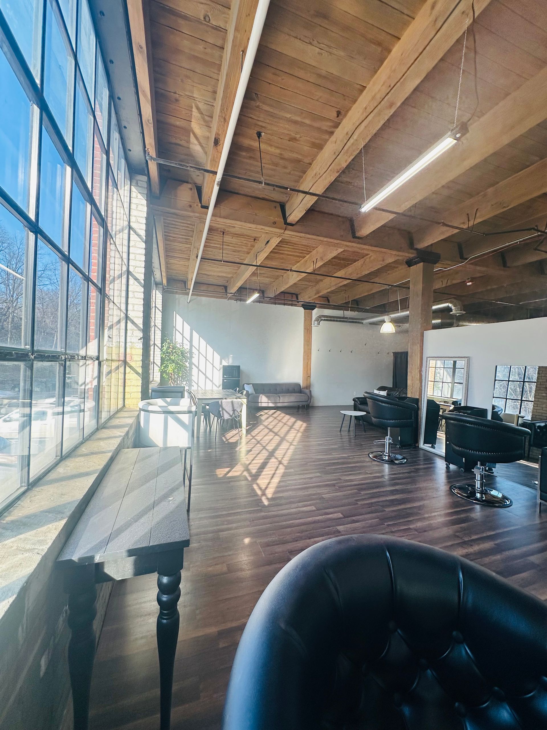 A sunlit salon with large windows, exposed wooden ceiling beams, dark wood flooring, styling chairs, and workstations.