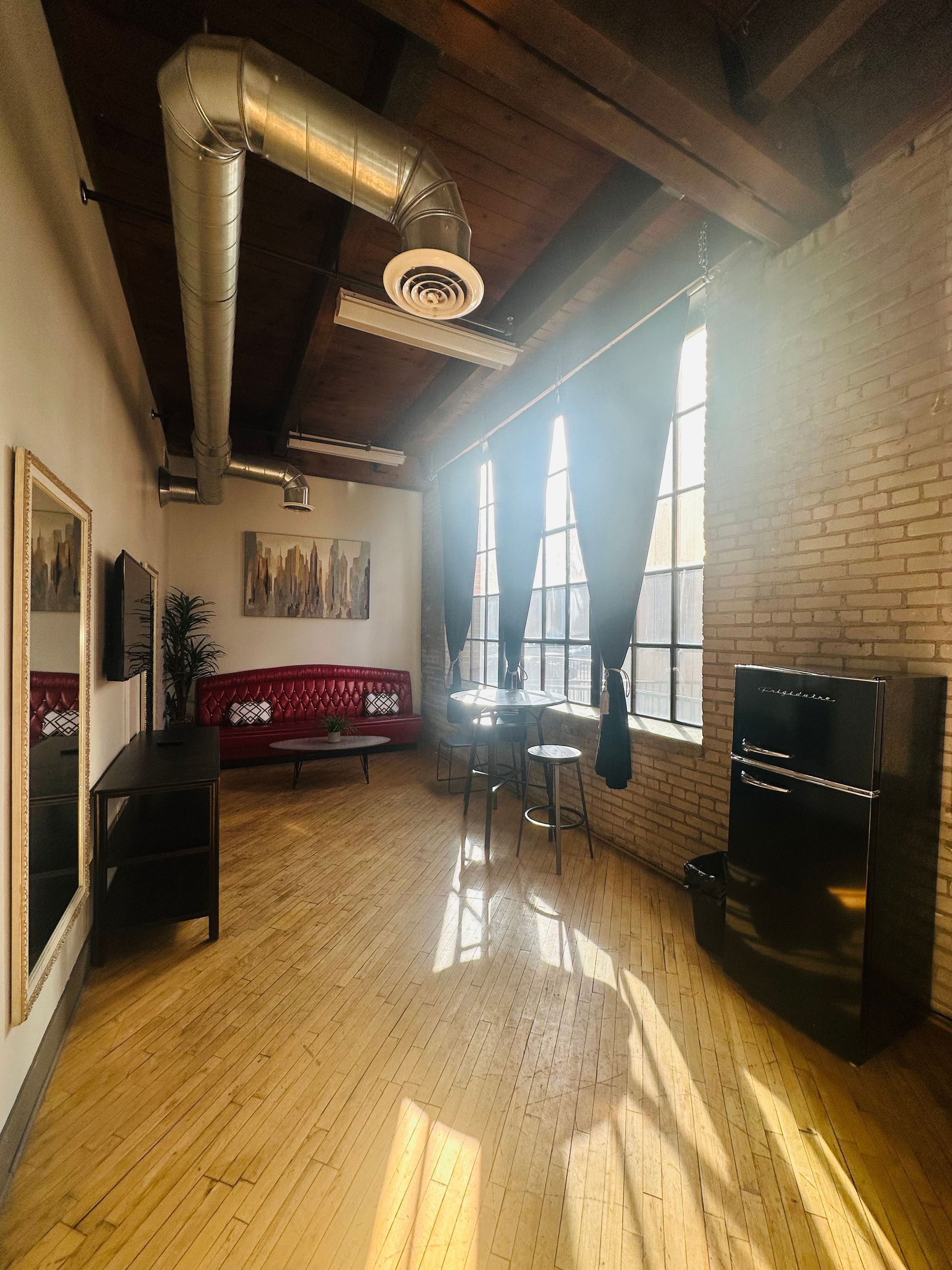 A loft-style room with a red leather sofa, wooden floor, exposed ceiling ducts, brick walls, and tall windows with drapes.