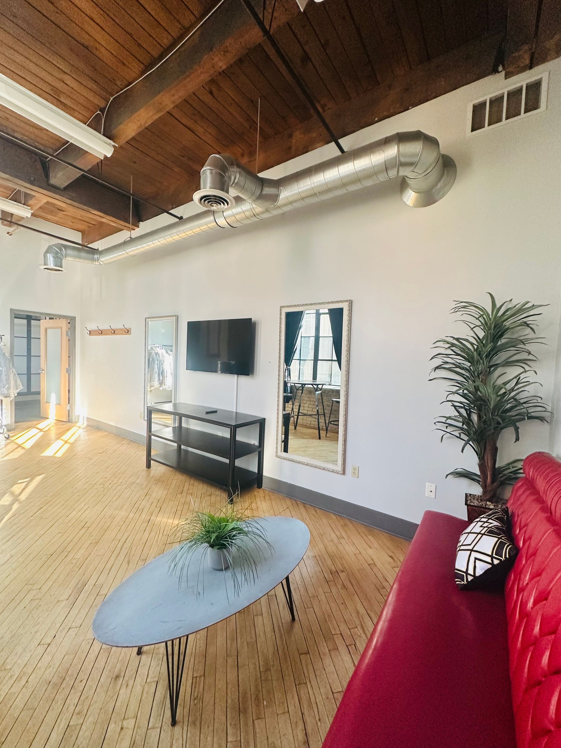 A bright loft living area with wood ceilings, an oval coffee table, a red sofa, a mounted TV, and a tall indoor plant.