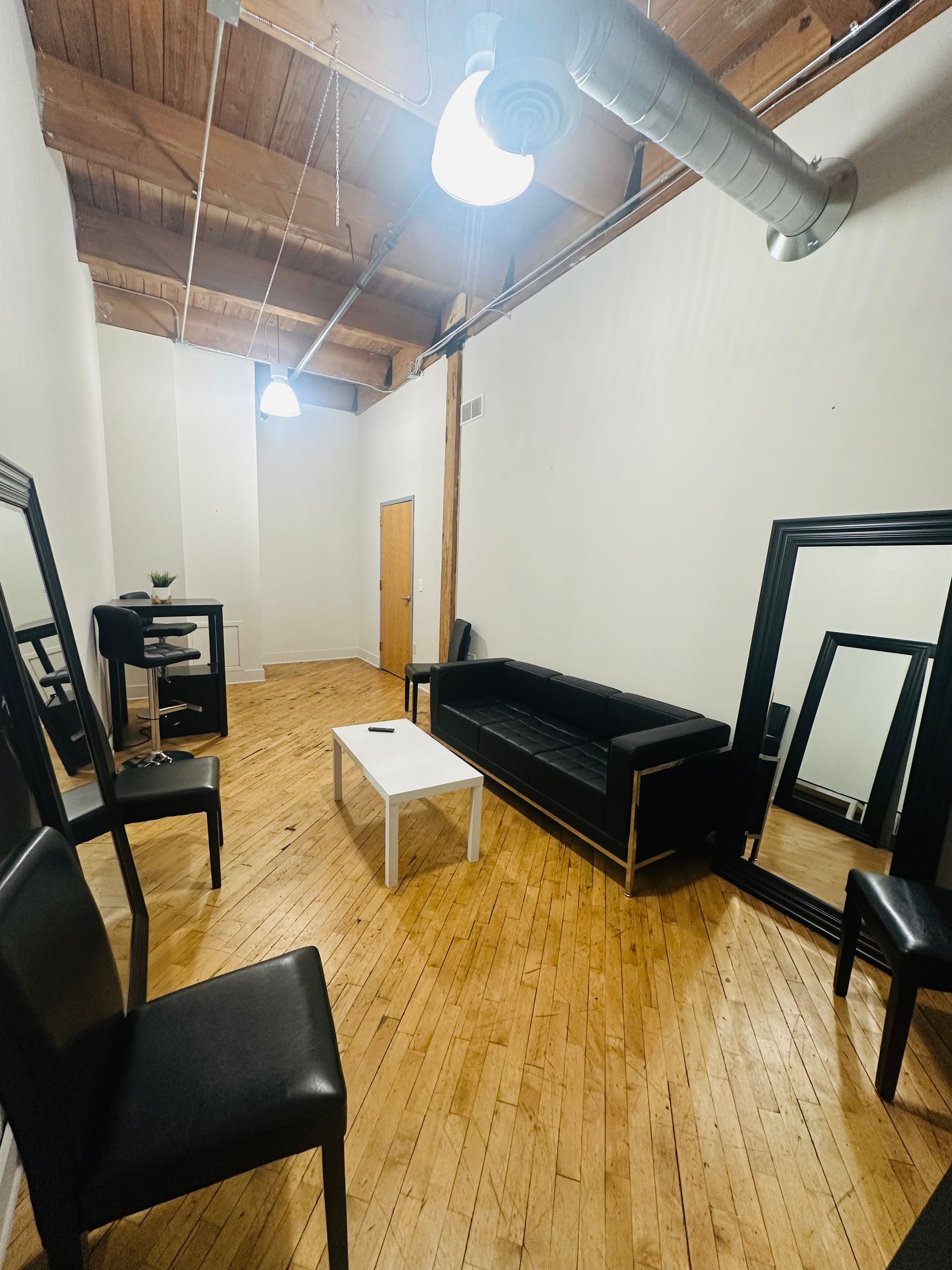 A room with wooden floors, a black sofa, a white coffee table, black chairs, and large mirrors against the white walls.