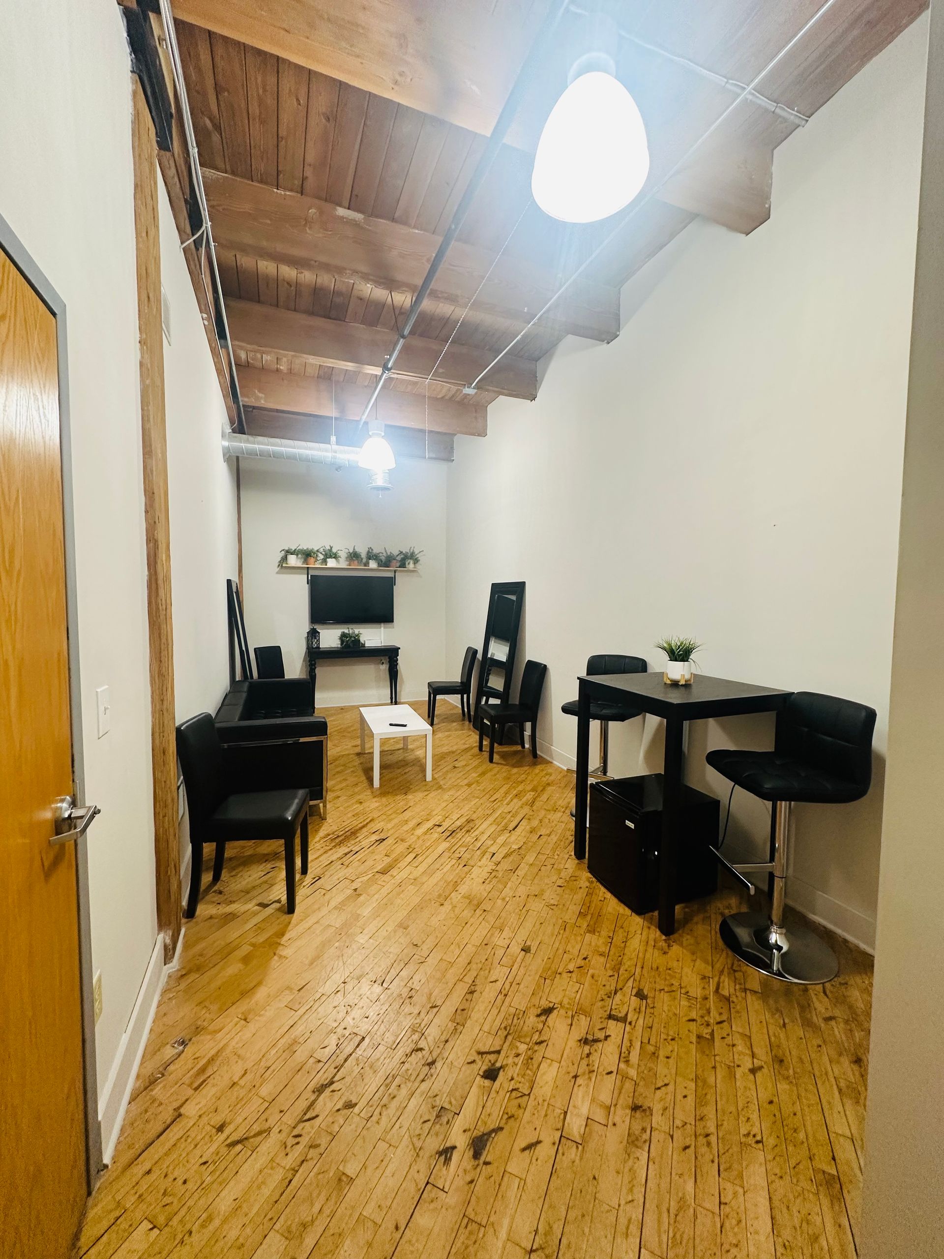 A narrow room with hardwood floors, a television, a small table with chairs, and an exposed wood beam ceiling.
