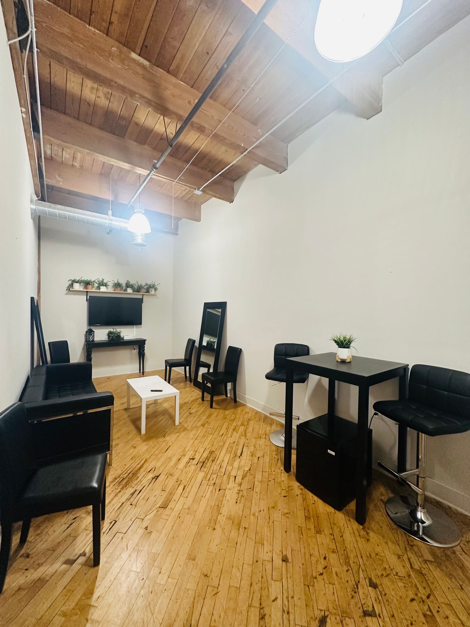 A long, narrow room with wooden floors, exposed ceiling beams, black furniture, and a wall-mounted TV.