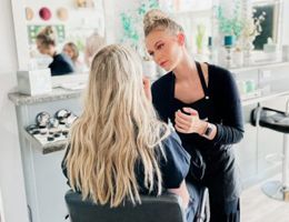 Woman applying makeup to another woman's face, salon setting, light-haired clients.
