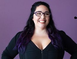Woman with glasses smiles, purple hair tips, black top, purple background.