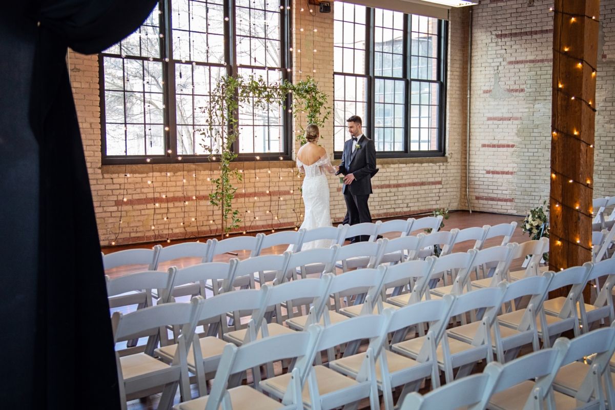 Bride and groom standing at the front of a rustic wedding venue with rows of white chairs