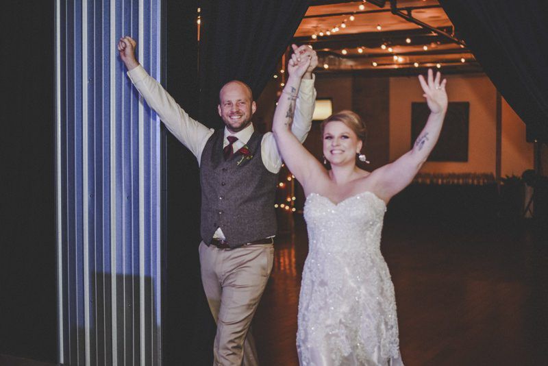 Newlyweds entering reception, arms raised in celebration, under warm string lights