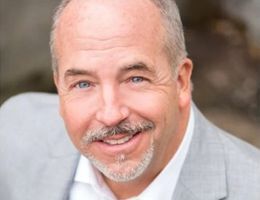Man in a gray suit smiles at the camera, light skin, short gray hair and goatee.