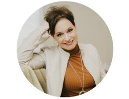 Woman smiling, wearing a light jacket and turtleneck, in a well-lit indoor setting.