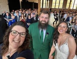 Woman taking selfie with bride in white dress and groom in green suit at a wedding. Guests in the background.