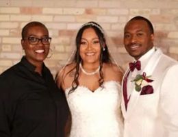 Wedding group: officiant in black, bride in white dress, groom in white suit, smiling.