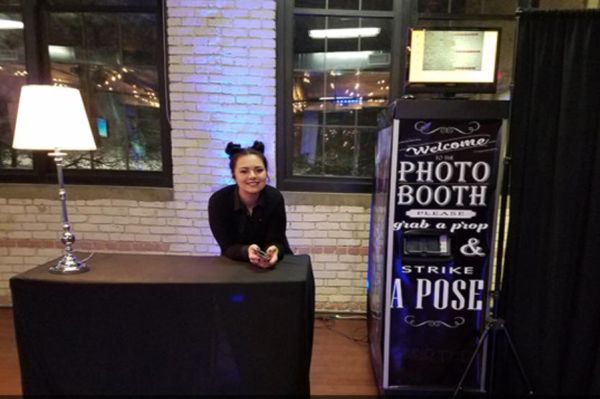 Woman with black cat-ear headband stands next to a photo booth and table with lamp.