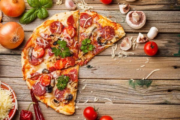 Pizza with toppings on a wooden table with ingredients like tomatoes, mushrooms, and herbs.