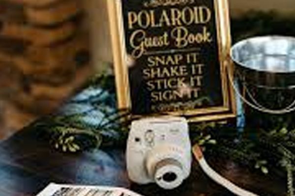 Polaroid guest book setup with camera, sign, bucket, and greenery.