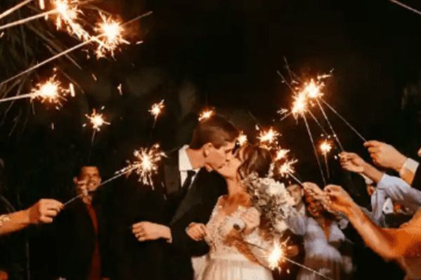Newlyweds kiss surrounded by guests holding sparklers, celebrating the couple outdoors at night.