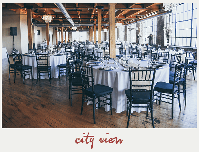 A large event space with round tables, chairs, and large windows. The wooden floor and ceiling give it an industrial vibe.