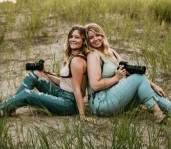Two women with cameras, sitting back-to-back in tall grass, smiling.