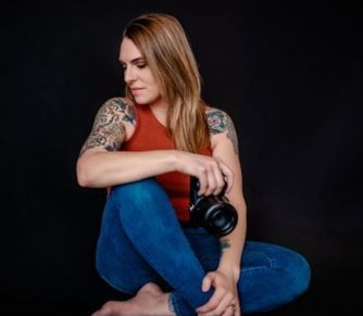 Woman with tattoos holding a camera, wearing a red top and blue jeans, sitting with a black background.