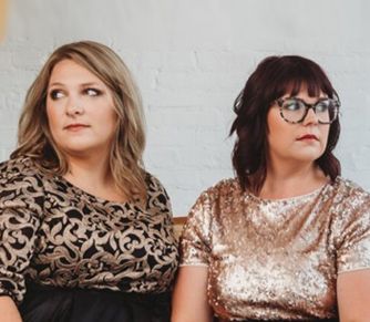Two women looking off-camera; one with blonde hair in black-and-gold top, one with dark hair in gold sequin top.