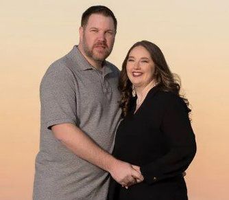 Man and woman holding hands, smiling, against a peach and yellow sky. The man wears a gray shirt. The woman wears a black top.