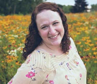 Woman smiling in a field of yellow and white flowers, wearing a floral blouse.