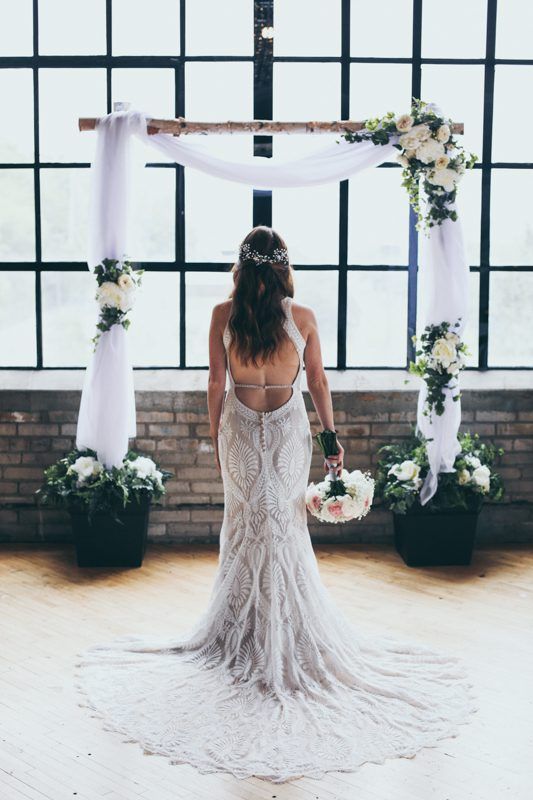 A bride in a beaded, backless wedding gown stands facing a large grid window under a flower-adorned wooden arch.