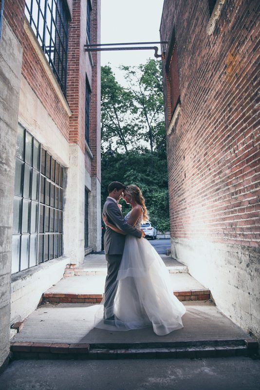 A couple in formal wedding attire embraces in a narrow alleyway between two brick buildings.