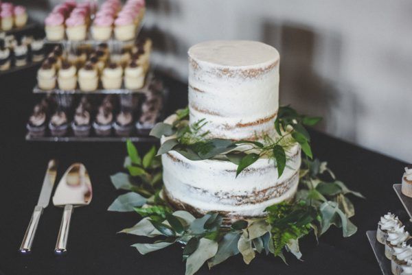 A two-tier semi-naked wedding cake decorated with greenery, next to a cupcake display and a cake serving set on a table.