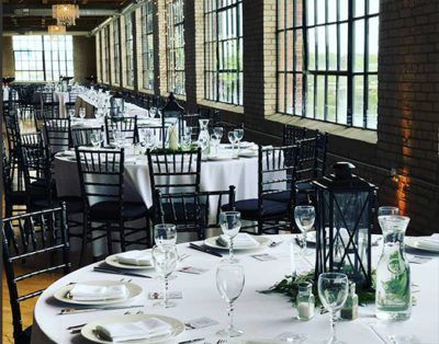 Tables set for a wedding reception in a venue with dark brick walls, large windows, and black chairs on white tablecloths.