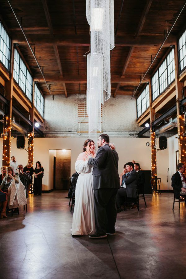 A couple dances in an industrial-style wedding venue with high ceilings, tall windows, and decorative hanging lights.