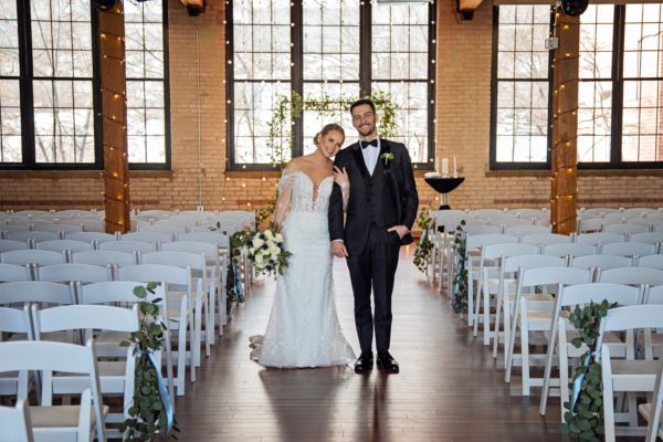 A couple poses together in the aisle of a sunlit indoor wedding venue filled with rows of empty white chairs.