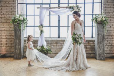 A person in a long lace wedding gown stands before a draped arch, while a child holds the end of the bridal veil.