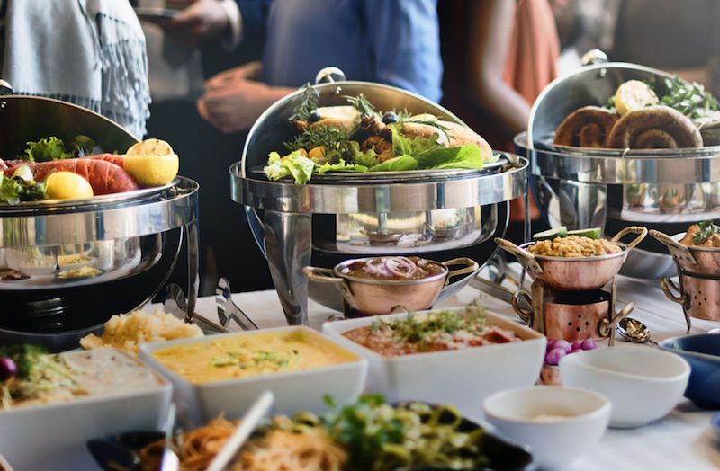 A buffet table featuring several silver chafing dishes filled with prepared foods and various side dishes in white bowls.
