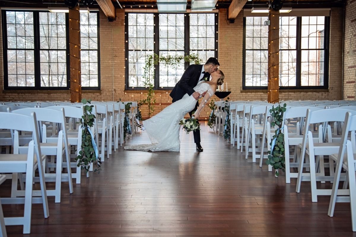 Couple kisses during a wedding ceremony in an industrial space, white chairs, and large windows.