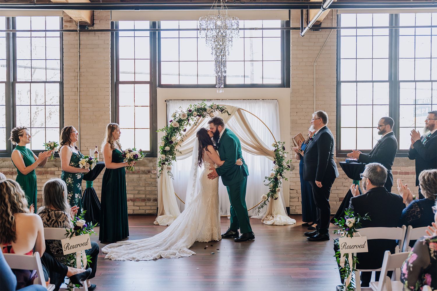 Wedding ceremony: couple kissing under floral arch, bridesmaids in green, guests clapping in a brick-walled venue.