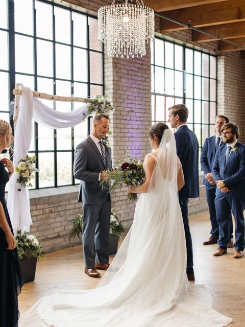 Wedding ceremony with couple, officiant, and groomsmen. Bride in white gown, groom in suit. Brick wall backdrop.