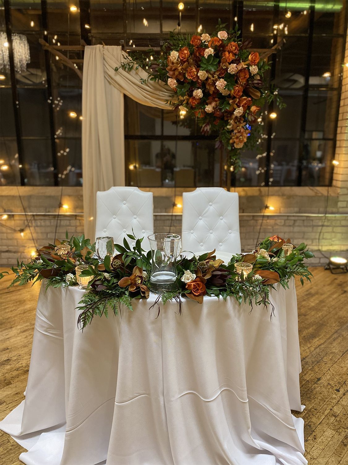 Wedding reception sweetheart table with floral arrangement, white chairs, and drapery, illuminated by string lights.