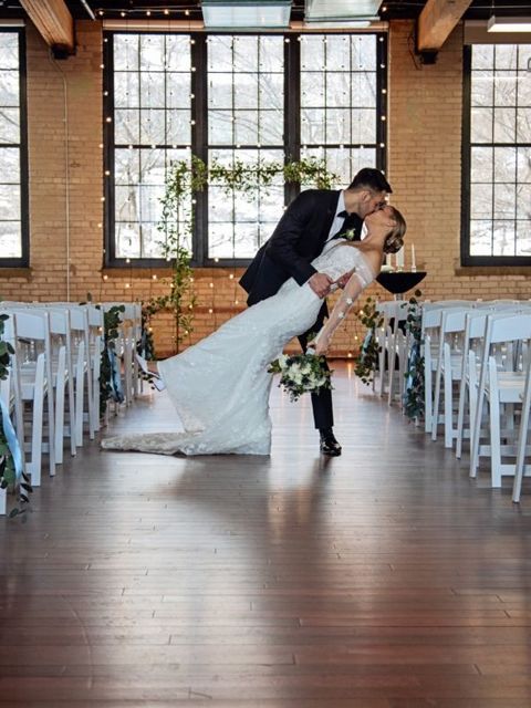 Groom dips bride for a kiss during wedding ceremony in a light-filled industrial space with white chairs and floral accents.