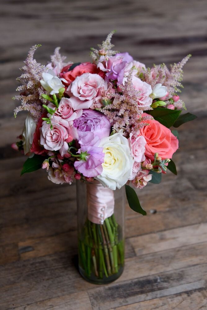 Bouquet of pink, peach, and purple flowers in a glass vase, tied with a pink ribbon, on a wooden surface.