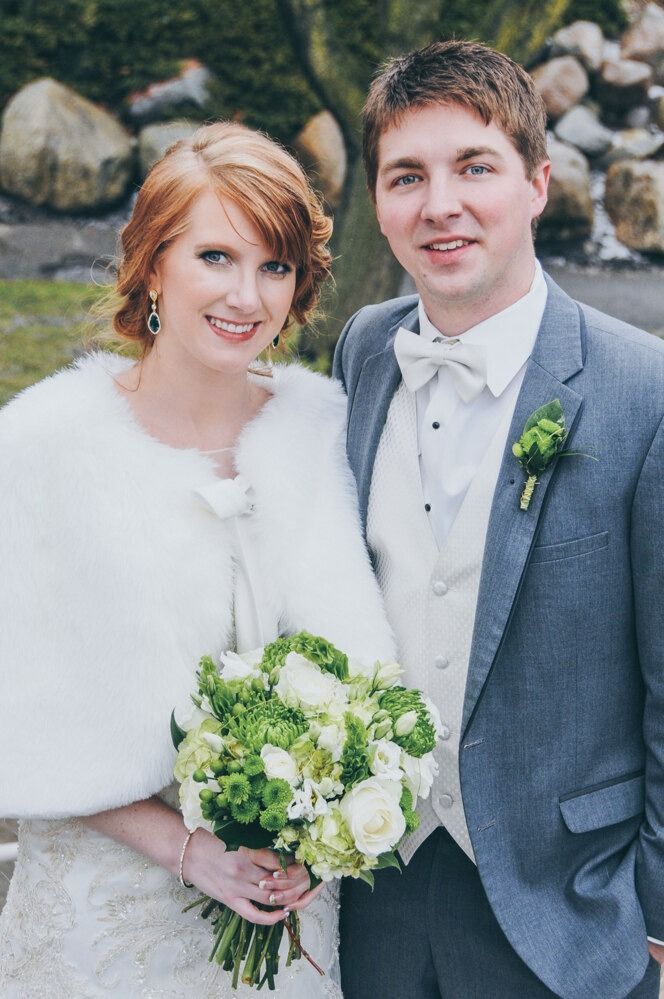 Bride and groom pose outdoors. She wears a white fur wrap, holds a bouquet. He wears a gray suit and bow tie.