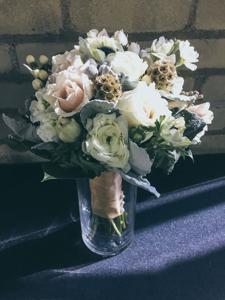 Bouquet of white, cream, and pink flowers in a glass vase, tied with tan ribbon, against a brick wall.