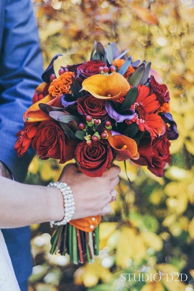 Bride and groom holding a vibrant autumn bouquet with red roses and orange calla lilies.