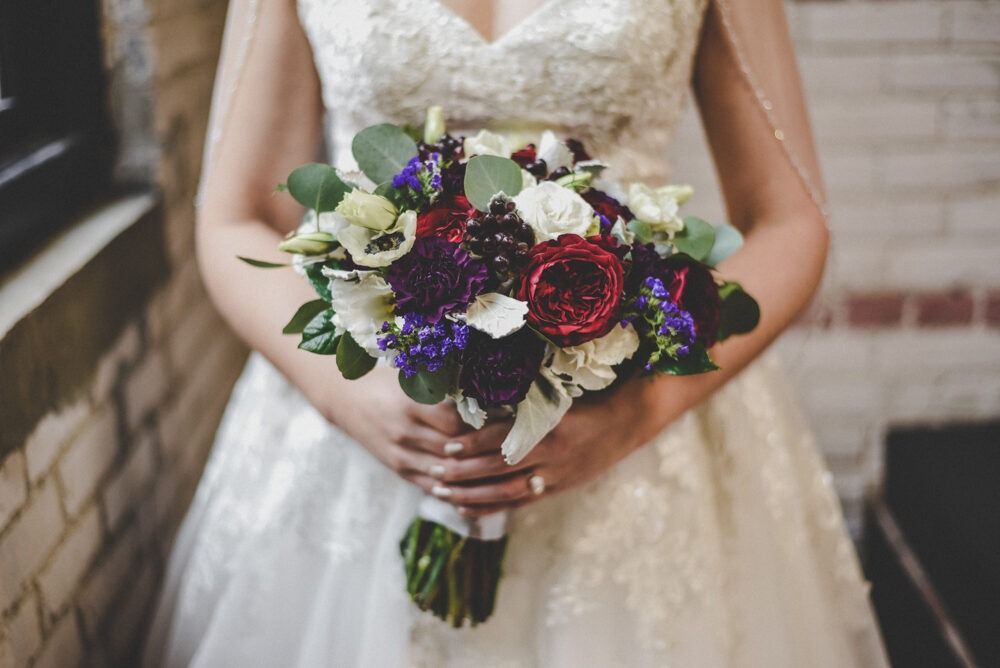 Bride in lace dress holding a bouquet of burgundy, purple, and white flowers.
