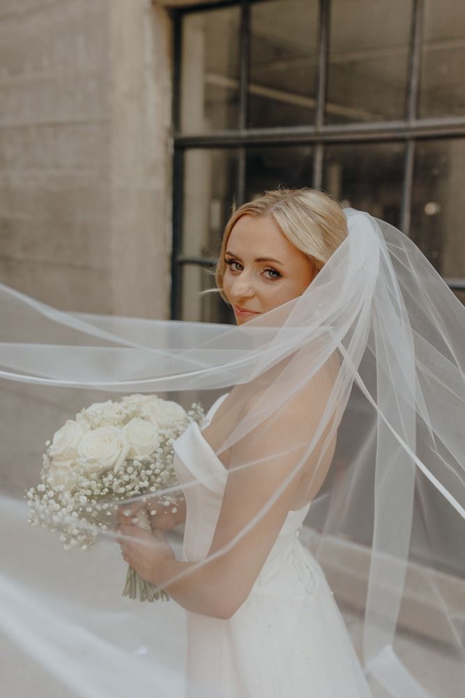 Bride holding a bouquet, covered in veil, smiling near a window.