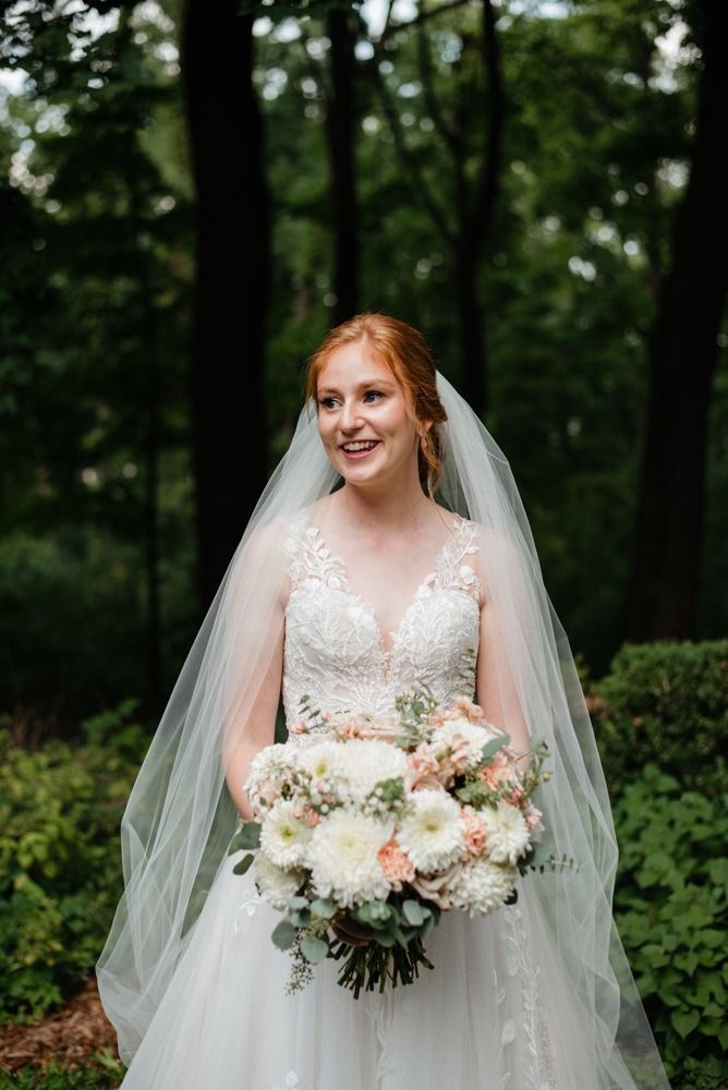 Bride holding a bouquet, wearing a white wedding dress and veil, smiling, surrounded by trees.