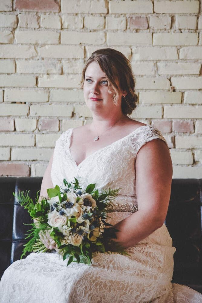Bride in lace dress holding bouquet, seated in front of a brick wall.