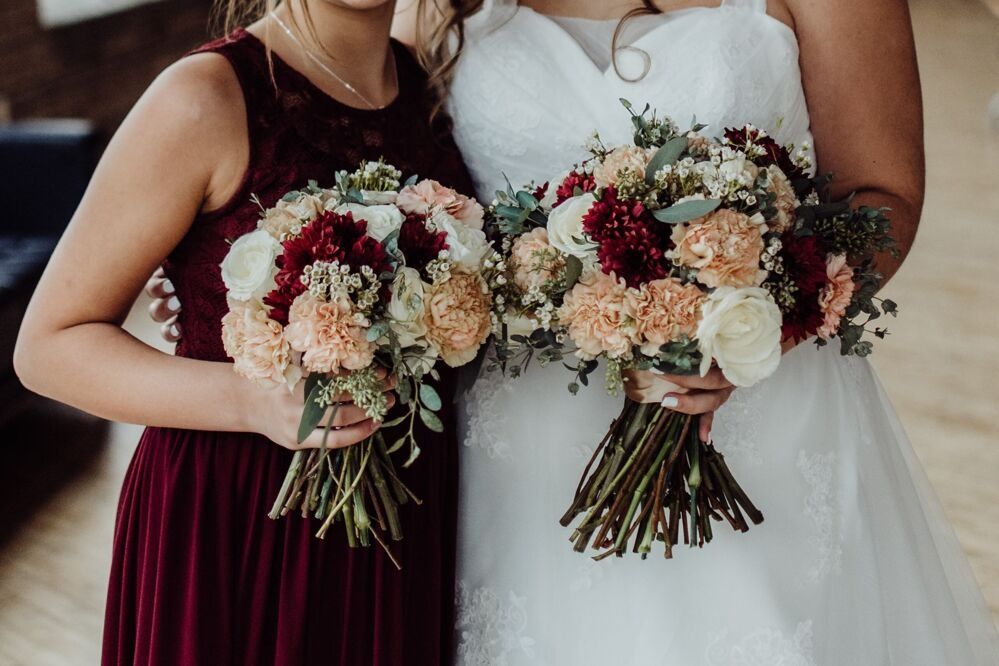 Bride and bridesmaid holding bouquets with red, peach, and white flowers; bridesmaid wears burgundy dress.
