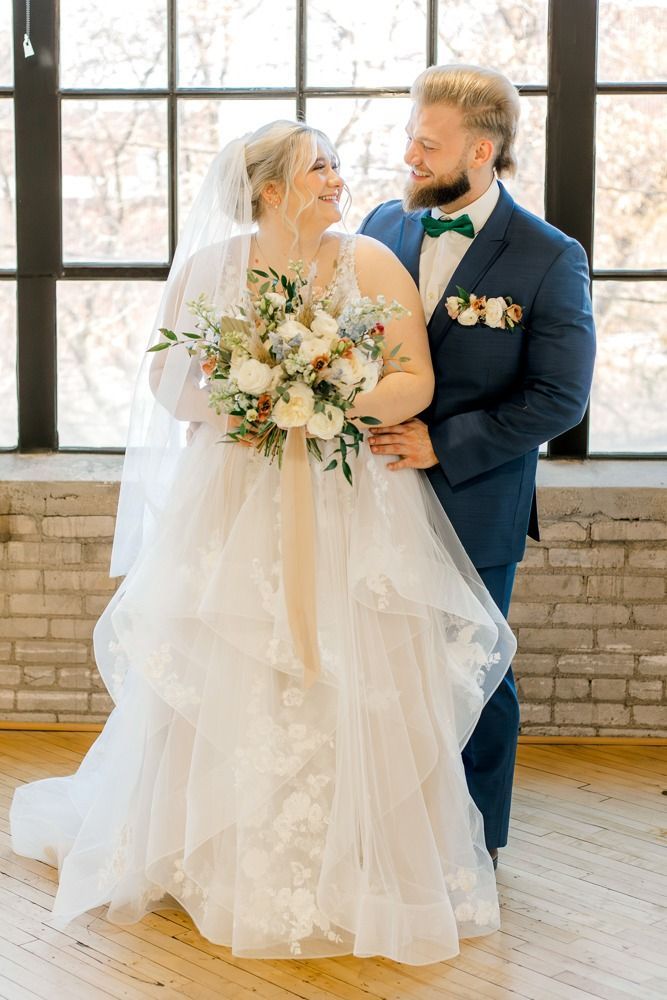 Bride and groom smiling at each other, holding hands. Bride in white dress and veil, groom in blue suit and green bow tie.