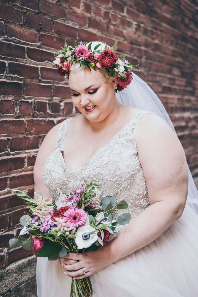 Bride in a white gown and floral crown, holding a bouquet, smiles near a brick wall.