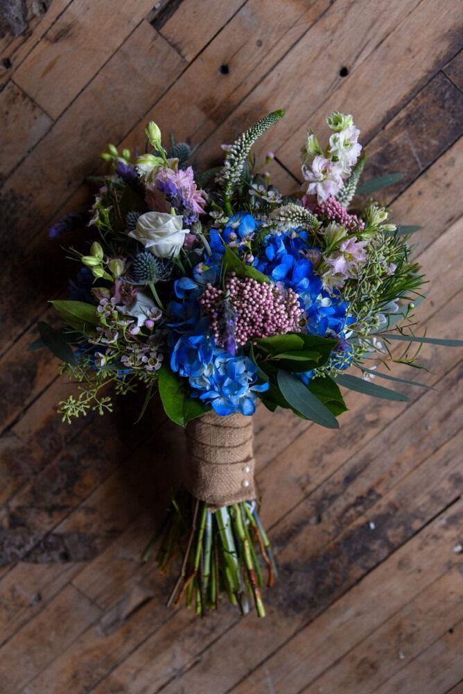 Bouquet of blue hydrangeas, thistle, and other flowers with a burlap-wrapped stem on a wood background.