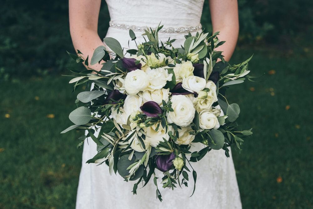 Bride in white dress holding a bouquet of white and purple flowers, greenery.